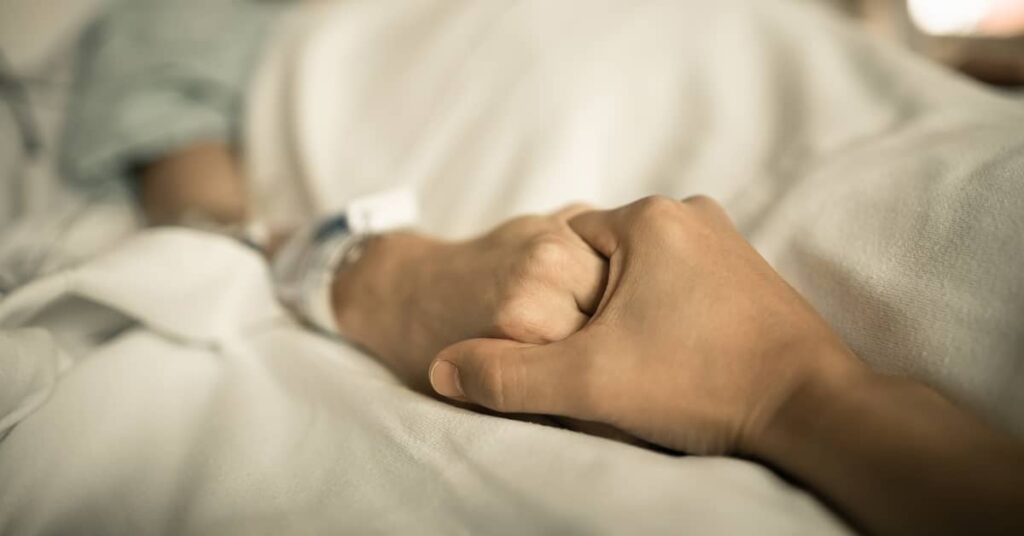 someone holds a patient's hand in a hospital bed | Burg Simpson