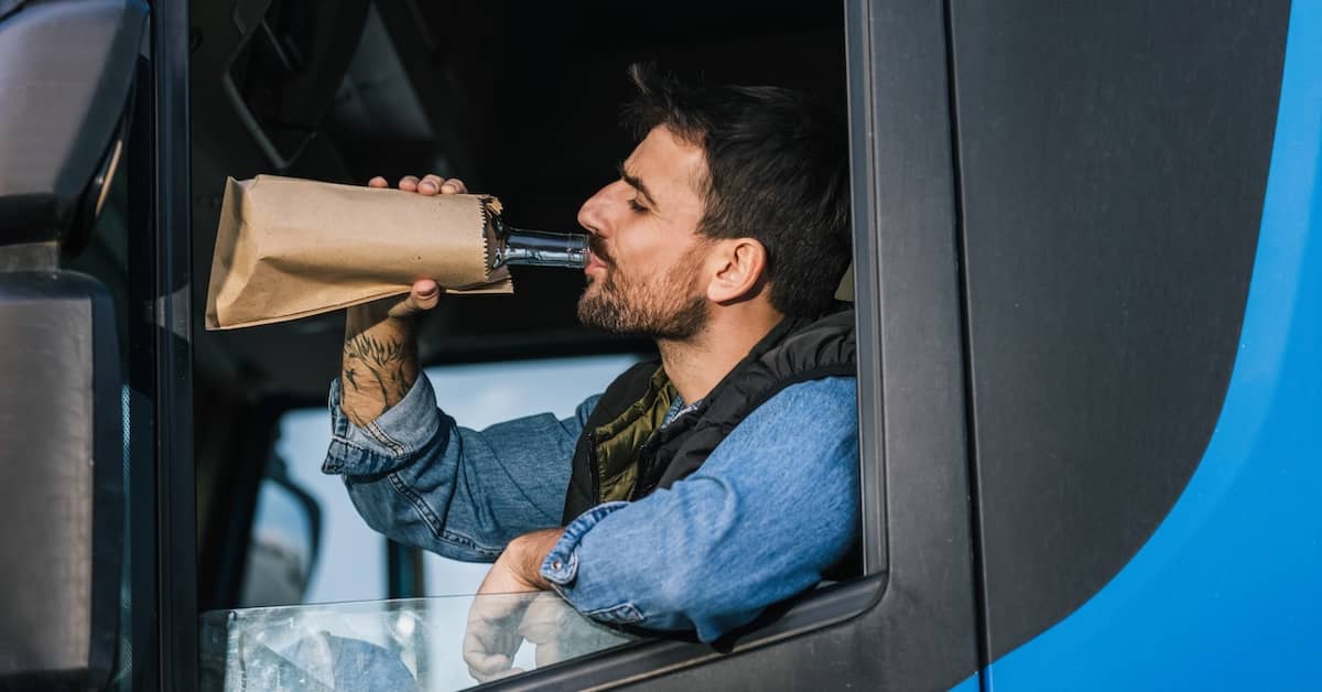 Truck driver drinking alcohol from a bottle in a brown paper bag in the cab of his rig | Burg Simpson